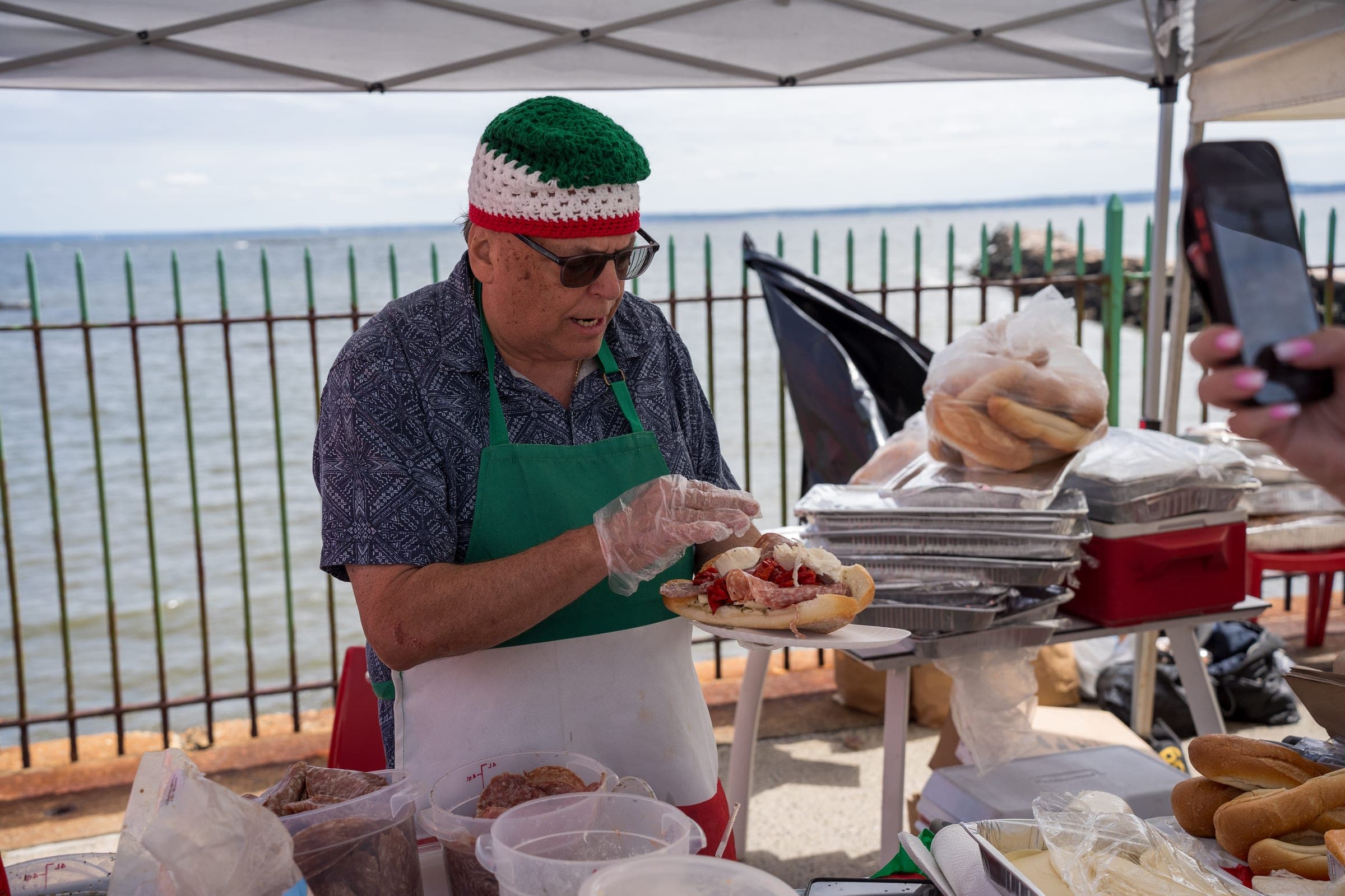 A vendor in a green, white and red cap builds a sausage and pepper hero at the pier rail, Long Island Sound behind him